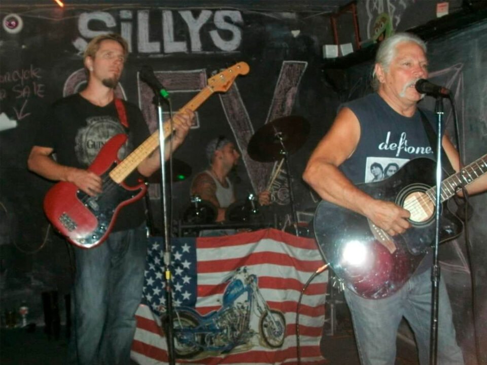 Cisco's Mexican Restaurant: Three musicians perform in a dimly lit venue. The guitarist and bassist are at the front, with the drummer in the background. An American flag with a motorcycle design adorns the drum kit.