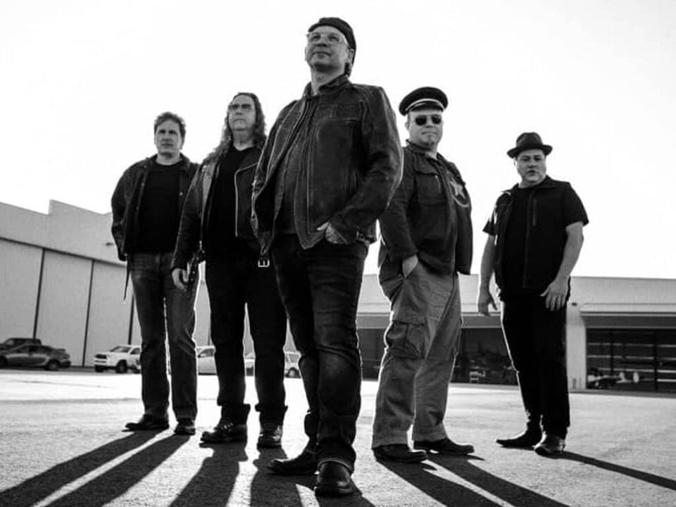 Cisco's Mexican Restaurant: Five men stand in a row outside in front of a large building, dressed in casual or military-inspired clothing, with a dramatic shadow cast behind them.